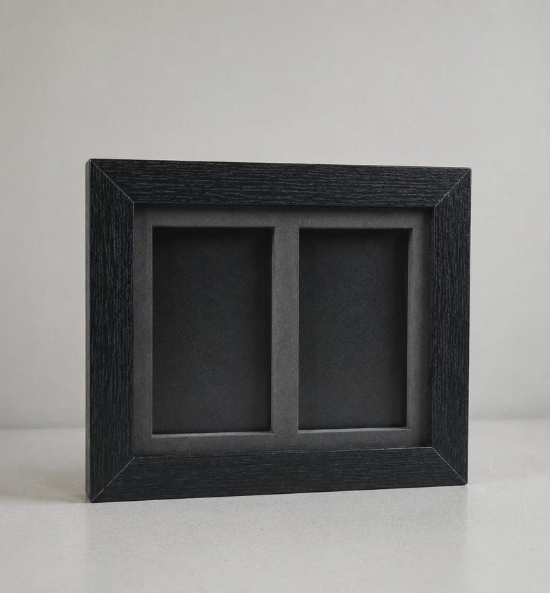 May include: A black, double-aperture picture frame. The frame has a textured wood grain appearance and a rectangular shape. The two openings are rectangular and have a dark gray background. The frame is designed to hold two photos.
