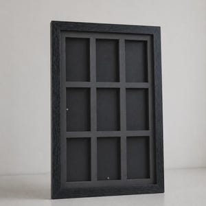 May include: A black rectangular picture frame with nine equally sized openings. The frame has a dark wood grain texture and is angled slightly, showcasing its depth and design. The interior is a solid black.