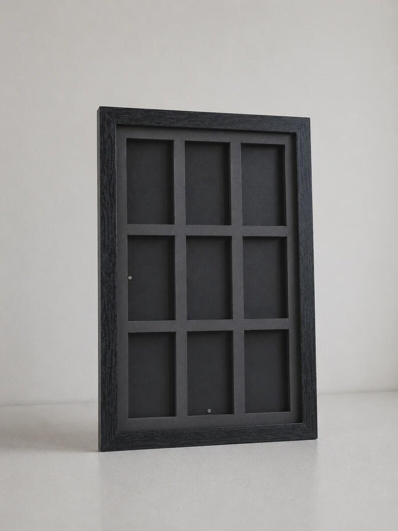 May include: A black rectangular picture frame with nine equally sized openings. The frame has a dark wood grain texture and is angled slightly, showcasing its depth and design. The interior is a solid black.