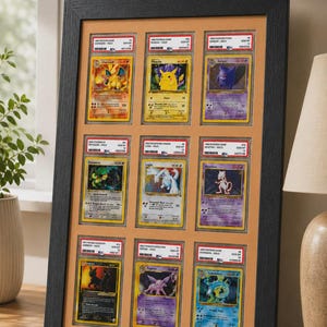 May include: A framed display of nine Pok&eacute;mon trading cards. The cards are in protective cases and arranged in a grid. The frame is black, and the background is a light orange color. The cards feature various Pok&eacute;mon characters.