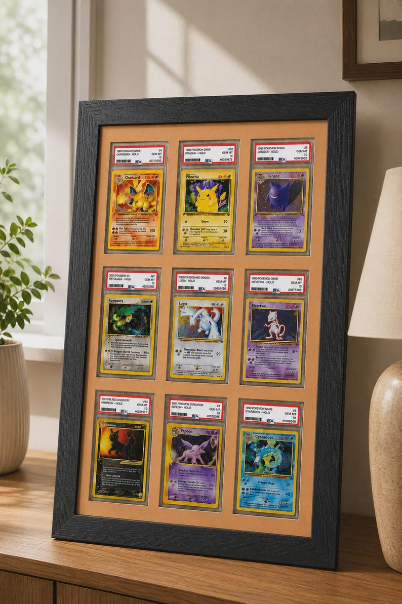 May include: A framed display of nine Pok&eacute;mon trading cards. The cards are in protective cases and arranged in a grid. The frame is black, and the background is a light orange color. The cards feature various Pok&eacute;mon characters.
