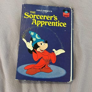 May include: A hardcover book titled Walt Disney's The Sorcerer's Apprentice with a dark blue cover. The front features a graphic of Mickey Mouse wearing a blue wizard hat with stars and a red robe, standing on a light blue oval shape.