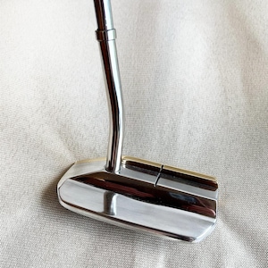 May include: A polished silver golf putter with a curved shaft and a rectangular head. The putter features a gold-coloured accent along the top edge. This club is designed for accuracy and control on the putting green.