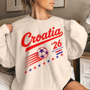 Puede incluir: Una sudadera de cuello redondo color crema con el texto en rojo Croatia, el n&uacute;mero 26 y las palabras SOCCER CLUB. El dise&ntilde;o incluye un gr&aacute;fico de un bal&oacute;n de f&uacute;tbol con l&iacute;neas de movimiento, una peque&ntilde;a bandera croata y una fila de estrellas rojas y azules.