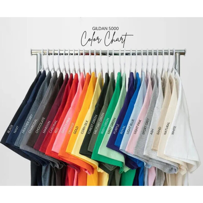 Puede incluir: Una carta de colores Gildan 5000 que muestra una fila de camisetas colgadas en perchas blancas. Las camisetas est&aacute;n dispuestas en un espectro de colores que incluye negro, azul marino, gris, marr&oacute;n, rojo, naranja, amarillo, verde, morado, azul, rosa y blanco.