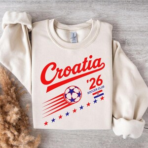 Puede incluir: Una sudadera de cuello redondo color crema con el texto en rojo Croatia, el n&uacute;mero 26 y las palabras SOCCER CLUB. Debajo hay un gr&aacute;fico de un bal&oacute;n de f&uacute;tbol con l&iacute;neas de movimiento, una peque&ntilde;a bandera croata y estrellas rojas y azules.