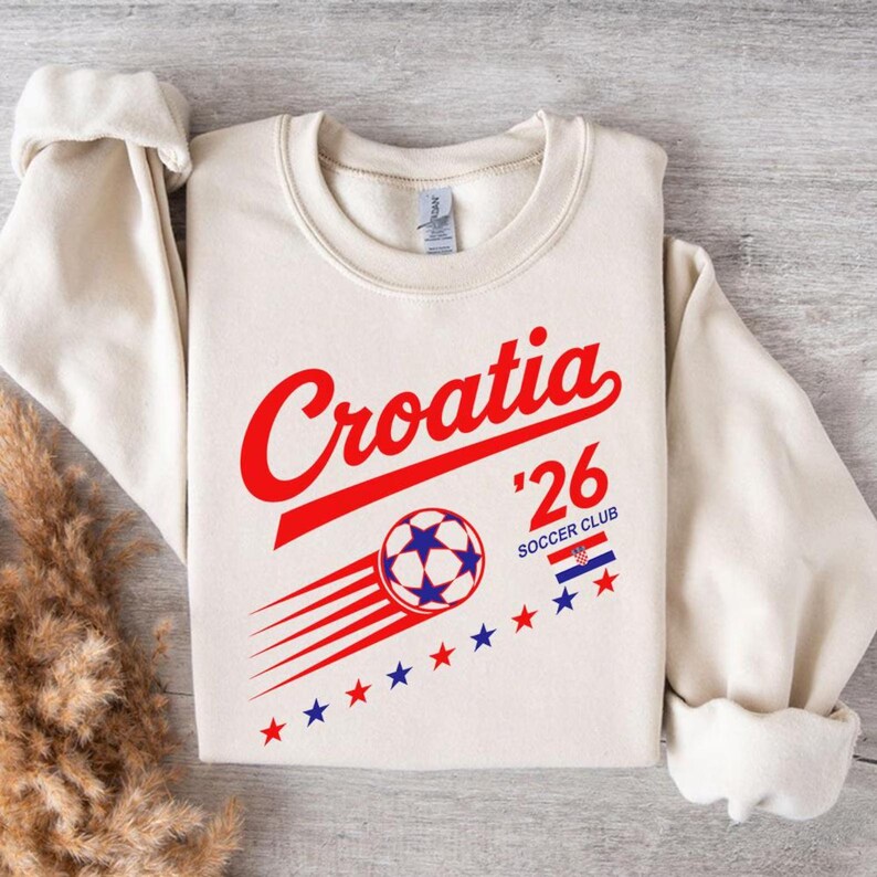 Puede incluir: Una sudadera de cuello redondo color crema con el texto en rojo Croatia, el n&uacute;mero 26 y las palabras SOCCER CLUB. Debajo hay un gr&aacute;fico de un bal&oacute;n de f&uacute;tbol con l&iacute;neas de movimiento, una peque&ntilde;a bandera croata y estrellas rojas y azules.