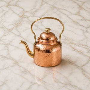 Puede incluir: Tetera de cobre con textura martillada, con asa, boquilla y tapa doradas. La tetera tiene un cuerpo redondeado y un pequeño pomo en la tapa. El diseño es tradicional y elegante.