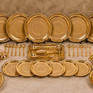 Puede incluir: Un gran juego de vajilla en tono dorado que incluye varios platos redondos, cuencos pequeños, vasos, cucharas, una bandeja rectangular y dos platos de servicio con tapa. Cada pieza presenta un intrincado diseño floral en relieve.