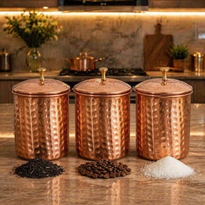 Könnte beinhalten: Drei kupferfarbene Vorratsdosen mit gehämmerten Strukturen und goldenen Knöpfen. Die zylindrischen Dosen haben Deckel und werden mit Teeblättern, Kaffeebohnen und Zucker präsentiert. Küchenaufbewahrung.