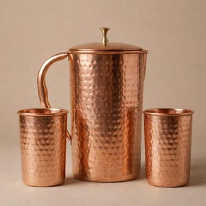 Peut inclure: Une carafe en cuivre avec un couvercle et une poignée, accompagnée de deux gobelets en cuivre assortis. L'ensemble a une texture martelée, lui donnant un aspect rustique et élégant. La carafe mesure environ 25 cm de haut.