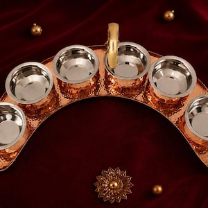 May include: A copper-colored serving tray with six stainless steel bowls arranged in a semi-circle. The tray has a hammered texture and a gold-colored handle. Decorative gold accents are scattered around the tray.