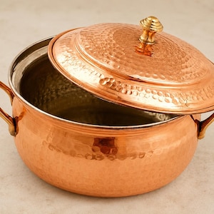May include: A copper cooking pot with a lid, featuring a hammered texture. The lid is slightly ajar, revealing the interior. The pot has two handles and a decorative knob on the lid. The pot is approximately 25 cm in diameter.