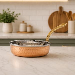 May include: Copper-coloured hammered saucepan with a gold-toned handle. The interior of the pan is silver. The pan is on a white countertop in a kitchen setting. Kitchenware.