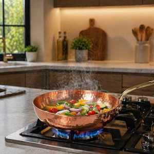 Puede incluir: Una sartén de color cobre llena de coloridas verduras, incluyendo tomates rojos, pimientos amarillos y cebollas rojas, se calienta en una estufa de gas. El vapor se eleva de la sartén, indicando que la comida se está cocinando. La cocina incluye gabinetes de madera.