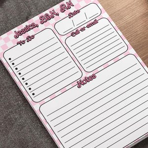May include: A pink and white checkered notepad with the text "Jessica, BSN, RN" at the top. The notepad has sections for "To-Do", "Date", "Call or email", and "Notes". The notepad is on a wooden desk.
