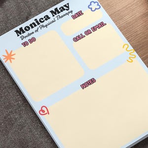 May include: A rectangular notepad with a light blue border and cream-coloured writing sections. The text reads Monica May Doctor of Physical Therapy, Date, Call or Email, To Do, and Notes. It features small colourful icons of a flower, star, and heart.