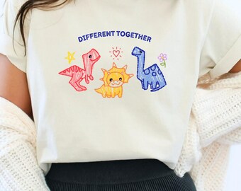 Può includere: T-shirt color avorio con la scritta "DIFFERENT TOGETHER" sopra tre dinosauri dei cartoni animati. I dinosauri sono rosa, gialli e blu, con una stella, un cuore e un fiore. La maglietta è a girocollo e maniche corte.