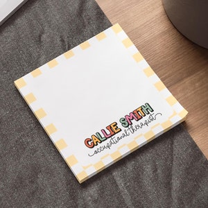 May include: A square notepad with a white center and a yellow and white checkered border. The text CALLIE SMITH is printed in colorful bubble letters, with the words occupational therapist written in black cursive script underneath the name.