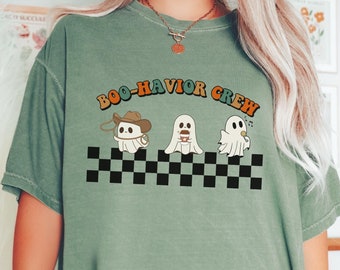 May include: Moss green t-shirt with the text "BOO-HAVIOR CREW" above a checkered pattern. The graphic features three cartoon ghosts, one wearing a cowboy hat, one with a coffee cup, and one singing. The word "MOSS" is printed on the bottom right.