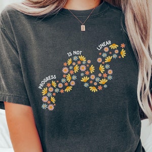 May include: A dark gray t-shirt with a floral design that reads "Progress is not linear".