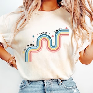 Pu&ograve; includere: Una maglietta bianca con un design grafico di una linea ondulata arcobaleno con il testo "Progress is not linear." in nero. La grafica &egrave; circondata da piccole stelle bianche.