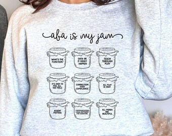 May include: Gray sweatshirt with the text "caba is my jam" printed on the front. The text is surrounded by nine illustrations of jars with different phrases inside each jar. The phrases are: "What's the function", "Data or it didn't happen", "Teach through play", "It's okay to feel all the feels", "Connection over compliance", "All play is okay", "Assent matters", "Communication looks different for everyone", "All minds are beautiful".