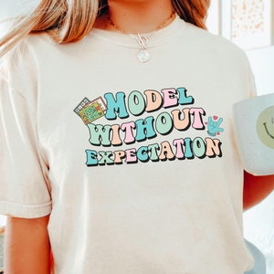 May include: A white t-shirt with the text "MODEL WITHOUT EXPECTATION" in colorful retro-style lettering. A small calculator graphic is above the text. The shirt is being worn by a person wearing light blue shorts.