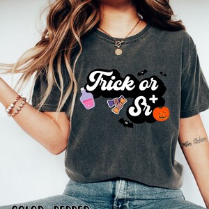 Puede incluir: Una camiseta gris oscuro con el texto "Trick or Sr+" en blanco y negro. La camiseta tiene un gr&aacute;fico de una botella de poci&oacute;n, caramelos, un murci&eacute;lago y una calabaza.