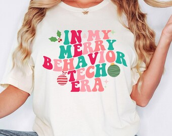 May include: A cream-colored short-sleeve t-shirt featuring the text "IN MY MERRY BEHAVIOR TECH ERA" in wavy, retro-style pink and teal lettering. The design includes small green holly leaves, a striped red ornament, and a green ornament.