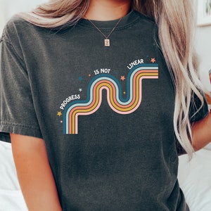 Puede incluir: Una camiseta gris oscuro con un diseño gráfico de una línea ondulada de colores arcoíris con estrellas y el texto "Progress is not linear".
