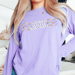 May include: A long-sleeved shirt in orchid purple with a graphic of a string of colorful beads spelling out "PHYSICAL THERAPY TECHNICIAN".