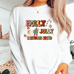 Op de afbeelding: Wit shirt met lange mouwen en een kleurrijke opdruk met de tekst "HOLLY JOLLY BEHAVIOR SQUAD". Het ontwerp bevat een sneeuwpop, kerstboom en zuurstok. Een casual, feestelijk kledingstuk.