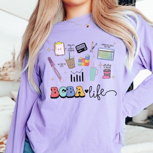 Puede incluir: Una camiseta de manga larga de color morado con el texto "BCBA-life" en letras de colores. La camiseta presenta una variedad de ilustraciones coloridas relacionadas con el campo del análisis del comportamiento, incluyendo un portapapeles, una gráfica, una taza de café y una calculadora.