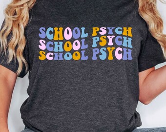 May include: A dark gray t-shirt with the text "SCHOOL PSYCH" repeated three times in a retro font. The text is in shades of pink, yellow, and blue.