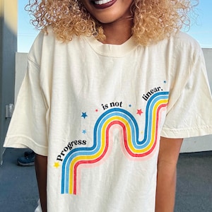 Pu&ograve; includere: Una maglietta color crema con una grafica di un arcobaleno con stelle e il testo "Progress is not linear."