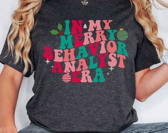 May include: A dark charcoal gray short-sleeve t-shirt featuring the text IN MY MERRY BEHAVIOR ANALYST ERA in wavy, multi-colored lettering. The design includes small green holly leaves, a green striped ornament, and a red striped ornament.