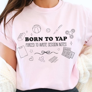 Puede incluir: Camiseta rosa claro con el texto negro "BORN TO YAP FORCED TO WRITE SESSION NOTES". El diseño incluye ilustraciones de estilo garabato, como una pelota, un bolígrafo y un fidget spinner. La palabra "blossom" está impresa en la esquina inferior derecha.