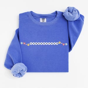 Puede incluir: Una sudadera azul con la palabra "SONOGRAPHER" escrita en cuentas blancas con acentos coloridos. La sudadera es de cuello redondo y tiene un ajuste holgado.
