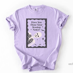 Op de afbeelding: Een lichtpaars t-shirt met korte mouwen en een donkere rechthoekige afbeelding met een geschulpte rand. De afbeelding toont een wit skelet, een kleine spin, sterren, vleermuizen en de tekst Have You Done Your Session Notes? in donkere letters.