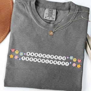 May include: Gray Comfort Colors t-shirt with the text "RADIOLOGIC TECHNOLOGIST" in white letter beads, decorated with colorful flowers, stars, and smiley faces. A gold necklace and a brown hat are also visible.
