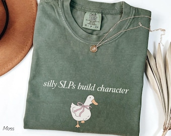 May include: A moss green t-shirt with the text "silly SLPs build character" and a cartoon duck wearing a pink bow. A gold necklace with a small pendant rests on the shirt. A brown hat and dried leaves are in the background.