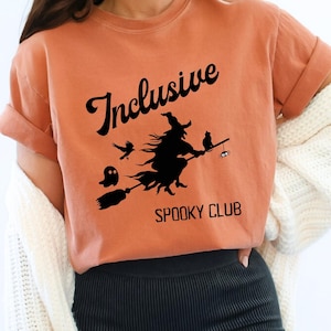 Puede incluir: Camiseta naranja con la frase "Inclusive Spooky Club" en negro y un diseño de una bruja en una escoba con un gato, un fantasma y una araña. Las mangas están dobladas.