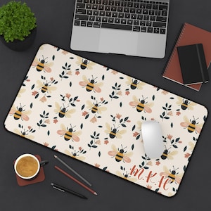 May include: A rectangular beige mouse mat with a black border, decorated with a pattern of yellow and black bees, pink flowers, and green leaves. The mat has the initials "m.k.ie" in red script.