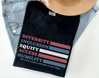 Può includere: T-shirt nera con le parole "DIVERSITÀ, INCLUSIONE, EQUITÀ, ACCESSO, UMILTÀ" in un design a pila con accenti rossi, bianchi e blu. Il testo "È così che costruiamo un mondo in cui tutti appartengono" è in basso. Un cappello beige e jeans blu sono sullo sfondo.