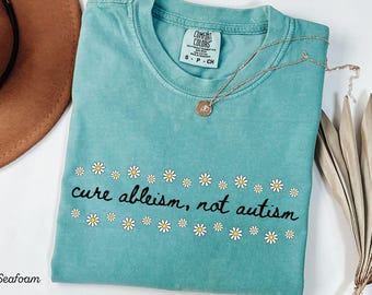 May include: Seafoam green t-shirt with the text "cure ableism, not autism" surrounded by small daisy graphics. A gold necklace with a pendant is draped across the shirt. The shirt is folded on a white surface.