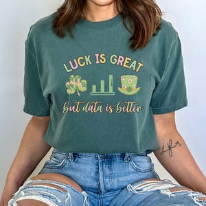 Könnte beinhalten: Ein grünes T-Shirt mit dem Text "Luck is great but data is better" in Regenbogenfarben. Das Shirt zeigt außerdem ein Kleeblatt, ein Balkendiagramm und einen Koboldhut.