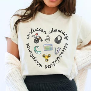 May include: Cream-colored t-shirt with a heart-shaped design featuring the words "inclusion, advocacy, acceptance, accessibility" and related icons. The icons include a wheelchair, a hand signing "I love you", headphones, a hearing aid, a calendar, a syringe, and a fidget spinner.