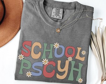 May include: A grey Comfort Colors t-shirt with the words "SCHOOL PSYCH" in colourful, retro-style lettering. White daisy graphics are interspersed throughout the text. A gold necklace with a pendant is draped across the shirt.