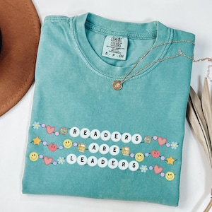 May include: A turquoise Comfort Colors t-shirt with the words "READERS ARE LEADERS" spelled out in white letter beads. The shirt is decorated with smiley faces, hearts, stars, and other colorful embellishments. A gold necklace rests on the shirt.